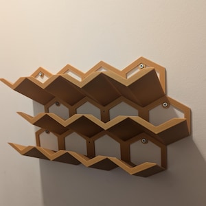 May include: A wall-mounted shelf unit made of wood with a honeycomb design. The shelf is painted a light brown color and has three tiers. The shelves are angled to create a unique look.