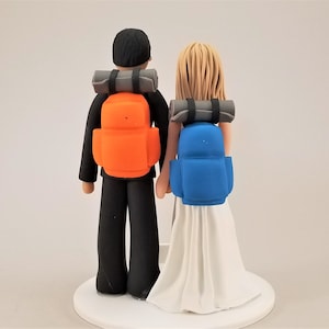 Op de afbeelding: Bruidstaarttopper met een bruid en bruidegom met rugzakken. De bruidegom heeft een oranje rugzak, de bruid een blauwe. Beide hebben grijze slaapmatten. De bruid draagt een witte jurk.