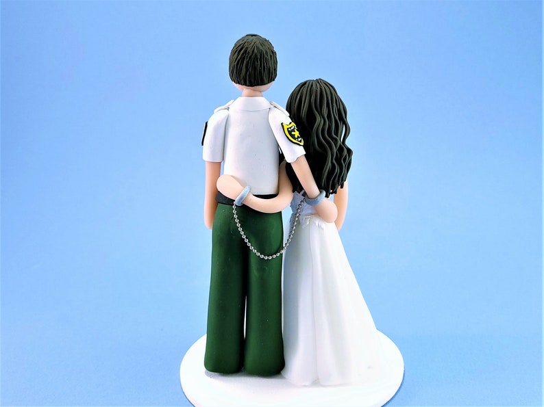 May include: A wedding cake topper featuring a couple in a playful pose. The groom is wearing a green uniform with a badge and the bride is wearing a white dress. They are connected by a silver chain.