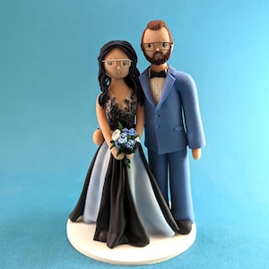 K&ouml;nnte beinhalten: Eine Tortenfigur f&uuml;r eine Hochzeit mit einer Braut- und Br&auml;utigamfigur. Die Braut tr&auml;gt ein schwarzes und hellblaues Kleid und h&auml;lt einen Blumenstrau&szlig;. Der Br&auml;utigam tr&auml;gt einen hellblauen Anzug mit einer schwarzen Fliege. Beide tragen eine Brille.