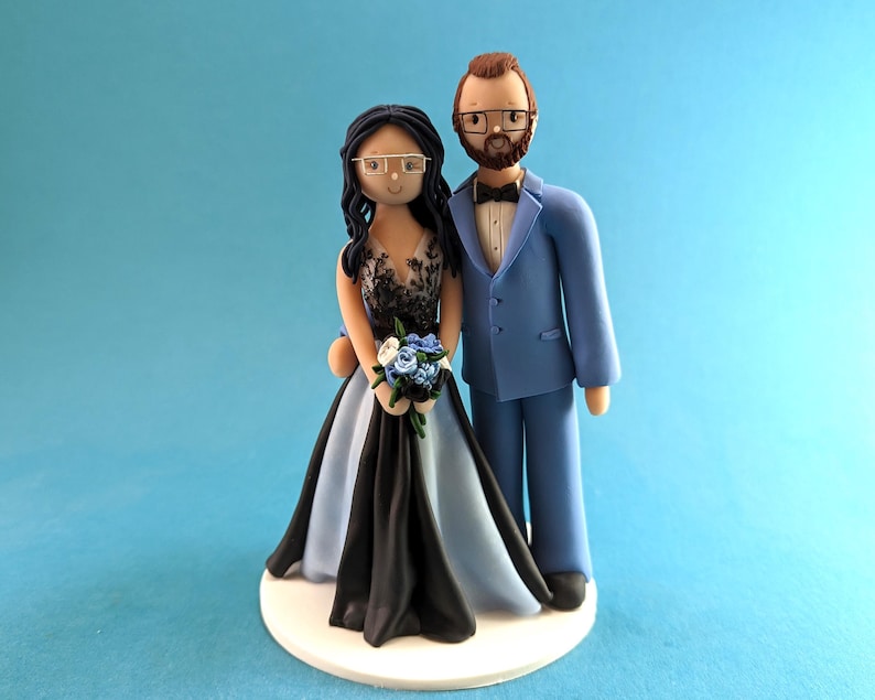 K&ouml;nnte beinhalten: Eine Tortenfigur f&uuml;r eine Hochzeit mit einer Braut- und Br&auml;utigamfigur. Die Braut tr&auml;gt ein schwarzes und hellblaues Kleid und h&auml;lt einen Blumenstrau&szlig;. Der Br&auml;utigam tr&auml;gt einen hellblauen Anzug mit einer schwarzen Fliege. Beide tragen eine Brille.