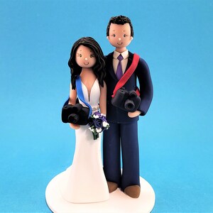 May include: A wedding cake topper featuring a bride and groom dressed in formal attire. The bride is wearing a white dress and a blue sash, while the groom is wearing a dark blue suit with a purple tie and a red camera strap. Both are holding cameras.