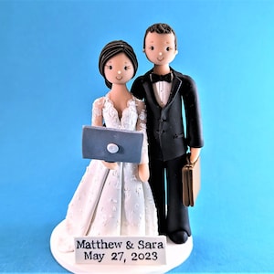 May include: A wedding cake topper featuring a bride and groom figurine. The bride is holding a laptop and the groom is holding a briefcase. The cake topper is on a white base with the text "Matthew & Sara May 27, 2023".