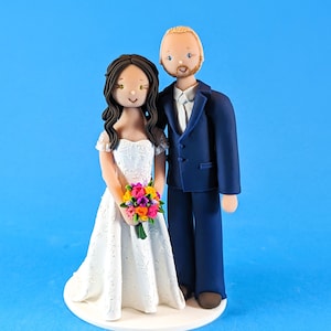 May include: A wedding cake topper featuring a bride and groom. The bride is wearing a white dress and holding a colorful bouquet. The groom is wearing a blue suit and a white shirt. The couple is standing on a white cake base.