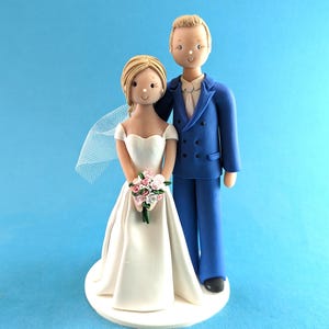 K&ouml;nnte beinhalten: Eine Tortenaufsatzfigur f&uuml;r eine Hochzeit, die ein Brautpaar zeigt. Die Braut tr&auml;gt ein wei&szlig;es Kleid und einen Schleier und h&auml;lt einen Strau&szlig; rosa und wei&szlig;er Blumen. Der Br&auml;utigam tr&auml;gt einen blauen Anzug. Die Figuren stehen auf einer wei&szlig;en Basis vor blauem Hintergrund.
