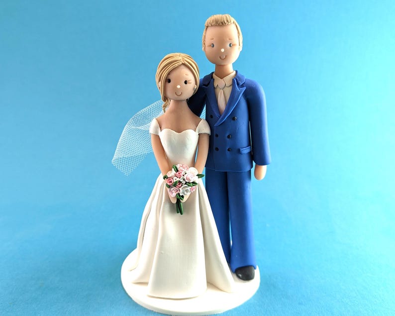 K&ouml;nnte beinhalten: Eine Tortenaufsatzfigur f&uuml;r eine Hochzeit, die ein Brautpaar zeigt. Die Braut tr&auml;gt ein wei&szlig;es Kleid und einen Schleier und h&auml;lt einen Strau&szlig; rosa und wei&szlig;er Blumen. Der Br&auml;utigam tr&auml;gt einen blauen Anzug. Die Figuren stehen auf einer wei&szlig;en Basis vor blauem Hintergrund.