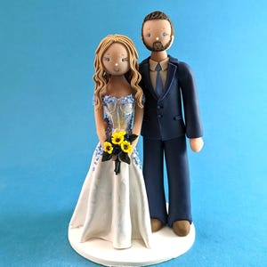 K&ouml;nnte beinhalten: Eine Tortenfigur f&uuml;r eine Hochzeit, die ein Brautpaar zeigt. Die Braut tr&auml;gt ein wei&szlig;es Kleid mit blauen Blumenakzenten und h&auml;lt einen Strau&szlig; gelber Blumen. Der Br&auml;utigam tr&auml;gt einen marineblauen Anzug. Die Figuren stehen auf einer wei&szlig;en Basis.