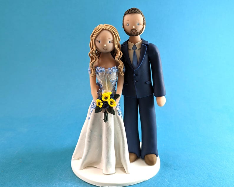 K&ouml;nnte beinhalten: Eine Tortenfigur f&uuml;r eine Hochzeit, die ein Brautpaar zeigt. Die Braut tr&auml;gt ein wei&szlig;es Kleid mit blauen Blumenakzenten und h&auml;lt einen Strau&szlig; gelber Blumen. Der Br&auml;utigam tr&auml;gt einen marineblauen Anzug. Die Figuren stehen auf einer wei&szlig;en Basis.
