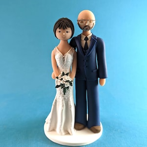 K&ouml;nnte beinhalten: Eine Tortenaufsatzfigur f&uuml;r eine Hochzeit, die eine Braut in einem wei&szlig;en Spitzenkleid mit einem Blumenstrau&szlig; und einen Br&auml;utigam in einem marineblauen Anzug zeigt. Die Figuren stehen auf einer wei&szlig;en Basis vor hellblauem Hintergrund.