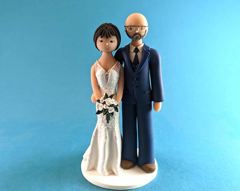 K&ouml;nnte beinhalten: Eine Tortenaufsatzfigur f&uuml;r eine Hochzeit, die eine Braut in einem wei&szlig;en Spitzenkleid mit einem Blumenstrau&szlig; und einen Br&auml;utigam in einem marineblauen Anzug zeigt. Die Figuren stehen auf einer wei&szlig;en Basis vor hellblauem Hintergrund.