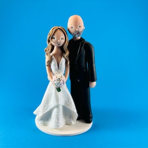 K&ouml;nnte beinhalten: Eine Tortenfigur f&uuml;r eine Hochzeit, die eine Braut in einem wei&szlig;en Kleid mit einem Blumenstrau&szlig; und einen Br&auml;utigam in einem schwarzen Anzug zeigt. Die Braut hat lange braune Haare und der Br&auml;utigam ist kahl mit Bart. Die Figuren stehen auf einer wei&szlig;en Basis vor blauem Hintergrund.