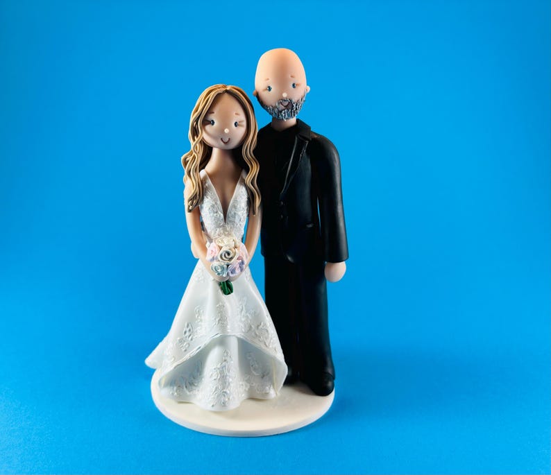 K&ouml;nnte beinhalten: Eine Tortenfigur f&uuml;r eine Hochzeit, die eine Braut in einem wei&szlig;en Kleid mit einem Blumenstrau&szlig; und einen Br&auml;utigam in einem schwarzen Anzug zeigt. Die Braut hat lange braune Haare und der Br&auml;utigam ist kahl mit Bart. Die Figuren stehen auf einer wei&szlig;en Basis vor blauem Hintergrund.