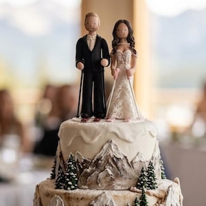 May include: A wedding cake with a mountain theme. The cake is decorated with a mountain range and miniature evergreen trees. A bride and groom cake topper is on top. The groom is wearing a black suit and the bride is wearing a white wedding dress.