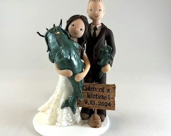May include: A wedding cake topper featuring a bride and groom holding catfish. The bride is holding a large catfish and the groom is holding a smaller catfish. The sign reads "Catch of a lifetime! 9.13.2024".