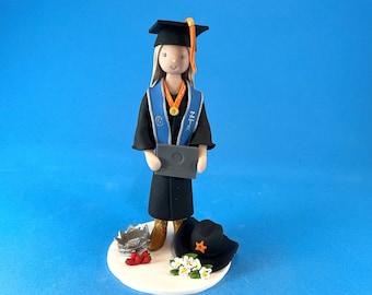 Könnte beinhalten: Eine Tortenfigur zur Graduierung, die eine Person in schwarzer Robe und Hut mit Diplom darstellt. Die Figur hat eine blaue Schärpe mit Text und eine goldene Medaille. Ein Cowboyhut, eine Krone und Blumen befinden sich am Sockel.
