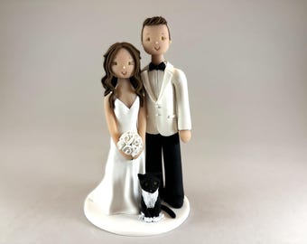 Pode incluir: Um topo de bolo de casamento branco com uma noiva e um noivo com um gato preto usando um colar branco e uma gravata borboleta.