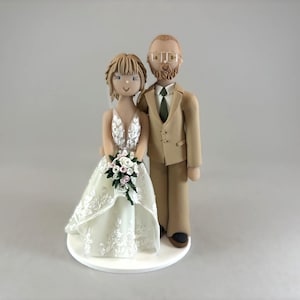 May include: A wedding cake topper featuring a bride and groom figurine. The bride wears a white, detailed wedding dress and holds a bouquet. The groom is in a tan suit with a green tie. The figurines stand on a white circular base.