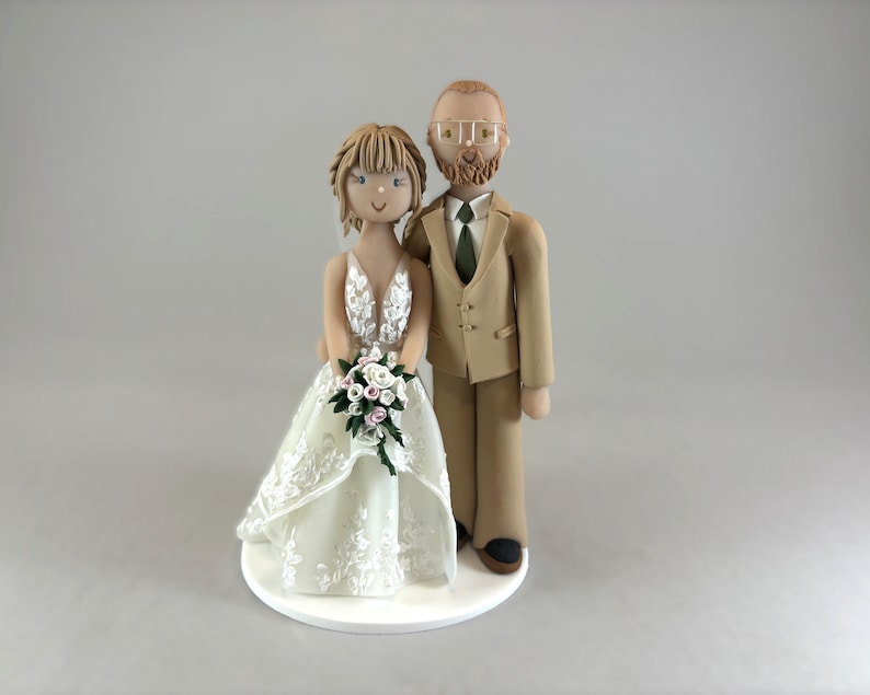 K&ouml;nnte beinhalten: Hochzeitstortenaufsatz mit einer Braut- und Br&auml;utigamfigur. Die Braut tr&auml;gt ein wei&szlig;es, detailliertes Hochzeitskleid und h&auml;lt einen Blumenstrau&szlig;. Der Br&auml;utigam tr&auml;gt einen beigefarbenen Anzug mit gr&uuml;ner Krawatte. Auf wei&szlig;er, runder Basis.
