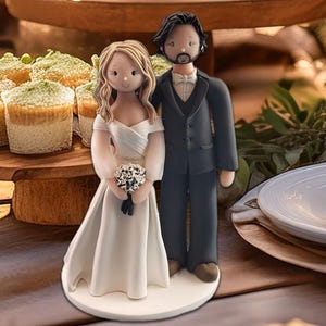 K&ouml;nnte beinhalten: Eine Tortenfigur f&uuml;r eine Hochzeit, die eine Braut in einem wei&szlig;en Kleid mit einem Blumenstrau&szlig; und einen Br&auml;utigam im schwarzen Anzug zeigt. Die Figuren stehen auf einer wei&szlig;en Basis. Cupcakes sind im Hintergrund zu sehen.
