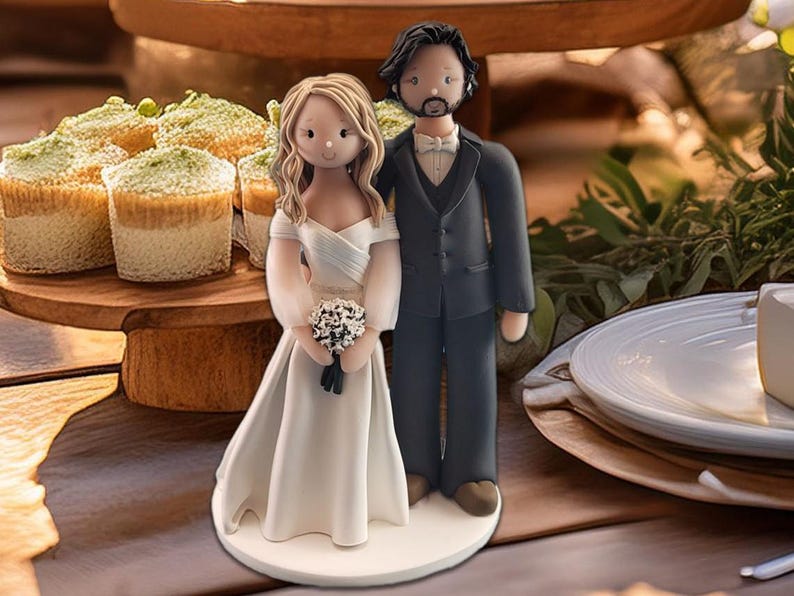 K&ouml;nnte beinhalten: Eine Tortenfigur f&uuml;r eine Hochzeit, die eine Braut in einem wei&szlig;en Kleid mit einem Blumenstrau&szlig; und einen Br&auml;utigam im schwarzen Anzug zeigt. Die Figuren stehen auf einer wei&szlig;en Basis. Cupcakes sind im Hintergrund zu sehen.