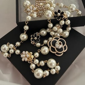 May include: A long, layered pearl necklace with black and white flower accents. The necklace features a gold charm with the number "5" in the center.