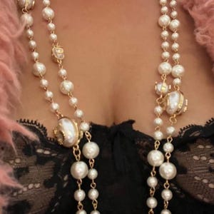 Pode incluir: Um colar de pérolas de duas voltas com detalhes dourados e grandes pingentes de pérolas. O colar é usado sobre uma blusa de renda preta.