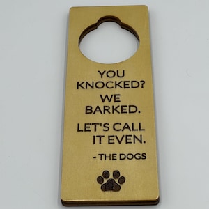 Puede incluir: Colgador de puerta rectangular de madera clara con un agujero circular en la parte superior. El texto "YOU KNOCKED? WE BARKED. LET'S CALL IT EVEN. - THE DOGS" está grabado en marrón oscuro. Una huella de pata en la parte inferior.
