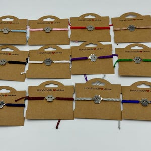 Op de afbeelding: Een verzameling handgemaakte vriendschapsarmbanden met een pootafdrukbedel. De armbanden worden gepresenteerd op bruine kartonnen labels en hebben verschillende koordkleuren, waaronder blauw, roze, rood en groen. Elk label heeft de tekst "Handmade with love."