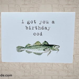 Może przedstawiać: Kartka okolicznościowa z napisem "i got you a birthday cod" nad akwarelową ilustracją ryby. Ryba jest zielono-biała z długą brodą na podbródku. Karta znajduje się na jasnym tle.