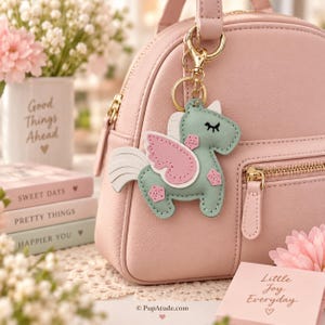 May include: A light pink backpack with a gold zipper and a mint green unicorn keychain featuring pink wings, a white mane, and a gold metal clasp. The unicorn has stitched details, including a closed eye and small pink pentagon shapes on its body.