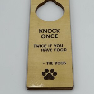 Puede incluir: Colgador de puerta de madera con un gran recorte circular en la parte superior. El texto "KNOCK ONCE TWICE IF YOU HAVE FOOD - THE DOGS" está impreso en negro. Una huella de pata está en la parte inferior.