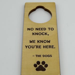 May include: Wooden door hanger with a decorative top and a large hole. The text reads "NO NEED TO KNOCK, WE KNOW YOU'RE HERE. - THE DOGS" with a paw print below. The wood is light brown.