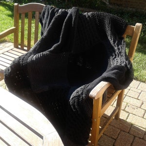 May include: A black knitted throw blanket draped over a wooden chair. The blanket has a textured pattern and is made of soft yarn.