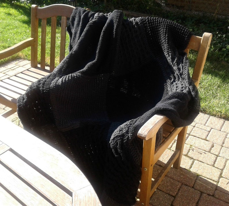 May include: A black knitted throw blanket draped over a wooden chair. The blanket has a textured pattern and is made of soft yarn.