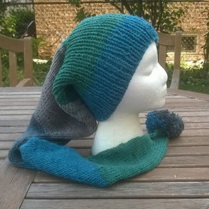 Puede incluir: Un gorro de punto azul, verde y gris con una larga bufanda adjunta. El gorro tiene una punta puntiaguda y la bufanda tiene un pomp&oacute;n en el extremo.