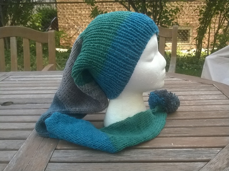 Puede incluir: Un gorro de punto azul, verde y gris con una larga bufanda adjunta. El gorro tiene una punta puntiaguda y la bufanda tiene un pomp&oacute;n en el extremo.