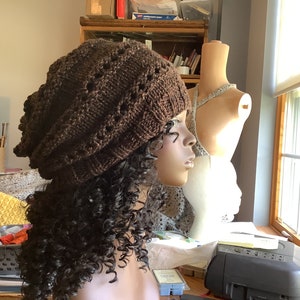 May include: A brown knitted beanie with a lacy pattern. The beanie is on a mannequin with curly brown hair.