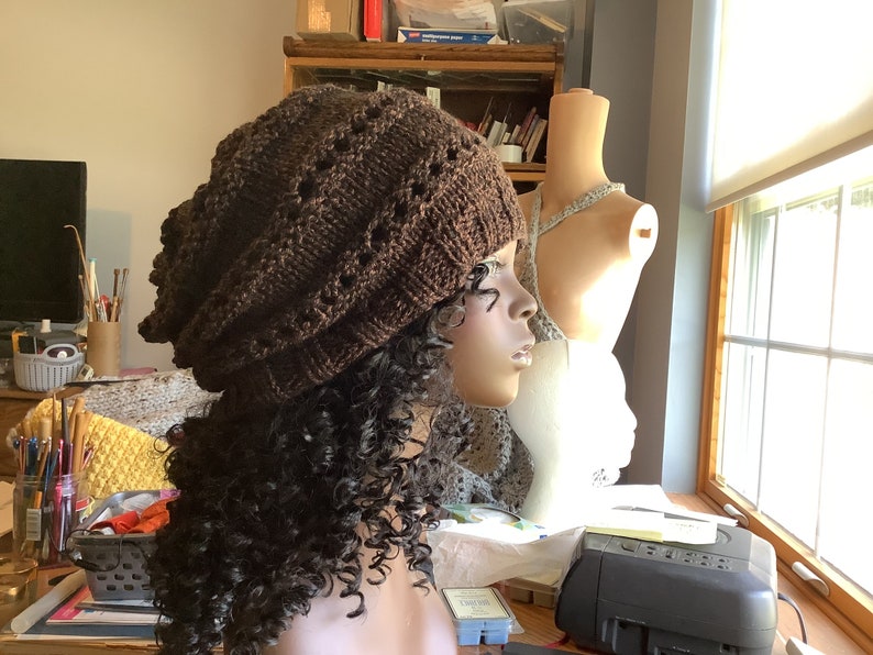 May include: A brown knitted beanie with a lacy pattern. The beanie is on a mannequin with curly brown hair.