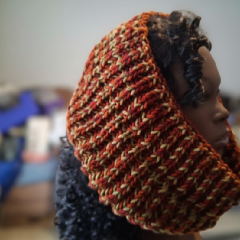 May include: A chunky knit cowl scarf featuring a ribbed texture with a variegated color pattern of deep burnt orange, rust red, and muted mustard yellow yarn. The accessory is worn around the neck and head, showcasing a thick, warm construction.
