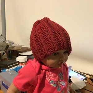 May include: A close-up of a child wearing a red knitted hat. The hat has a ribbed texture and is pulled down over the child's forehead. The child is wearing a pink shirt with a butterfly design.