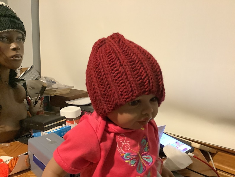 May include: A close-up of a child wearing a red knitted hat. The hat has a ribbed texture and is pulled down over the child's forehead. The child is wearing a pink shirt with a butterfly design.