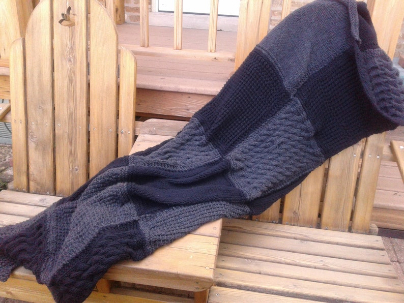 May include: A large, gray and black knitted throw blanket with a cable knit pattern. The blanket is draped over two wooden chairs.