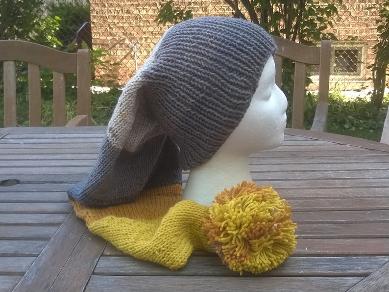 Puede incluir: Un gorro de punto gris y amarillo con una corona larga y puntiaguda y un gran pomp&oacute;n amarillo.