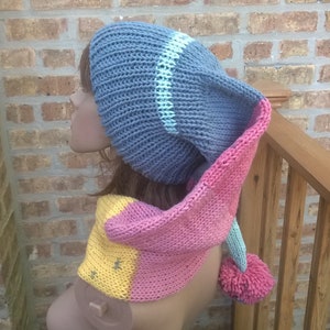 Puede incluir: Un gorro de punto azul y rosa con una larga bufanda adjunta. El gorro tiene una franja blanca y la bufanda tiene una franja amarilla. El gorro tiene un pomp&oacute;n en el extremo.