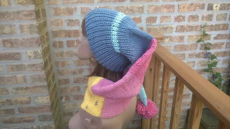 Puede incluir: Un gorro de punto azul y rosa con una larga bufanda adjunta. El gorro tiene una franja blanca y la bufanda tiene una franja amarilla. El gorro tiene un pomp&oacute;n en el extremo.