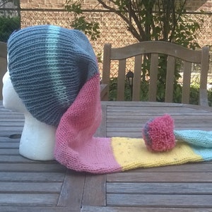 Puede incluir: Un gorro de punto gris, rosa, amarillo y azul con una cola larga. El gorro est&aacute; sobre una mesa de madera.