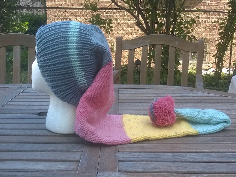 Puede incluir: Un gorro de punto gris, rosa, amarillo y azul con una cola larga. El gorro est&aacute; sobre una mesa de madera.