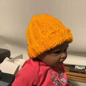 May include: A yellow knitted hat with a cable knit pattern. The hat is on a doll's head.