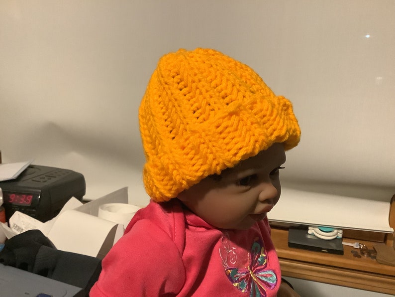 May include: A yellow knitted hat with a cable knit pattern. The hat is on a doll's head.