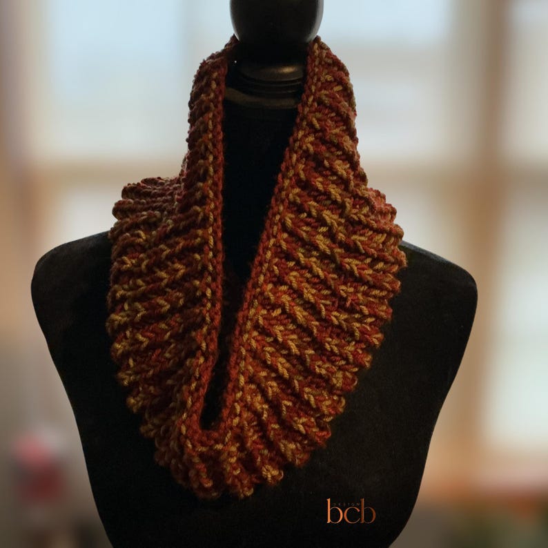 May include: A chunky knit cowl scarf featuring a ribbed texture with a variegated color pattern of deep rust red, burnt orange, and muted olive green yarn. The circular scarf is displayed on a black mannequin form with the text bcb at the bottom.